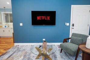 A living room with a TV mounted on a blue wall, a glass coffee table, a green armchair, and a small table with a lamp. - Chicago 3BR Oasis Near Downtown & United Center (Chicago)