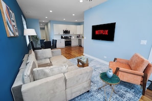 A living room with a sofa, armchair, and coffee table, a kitchen in the background, and a TV displaying 'NETFLIX'. - Chicago 3BR Oasis Near Downtown & United Center (Chicago)