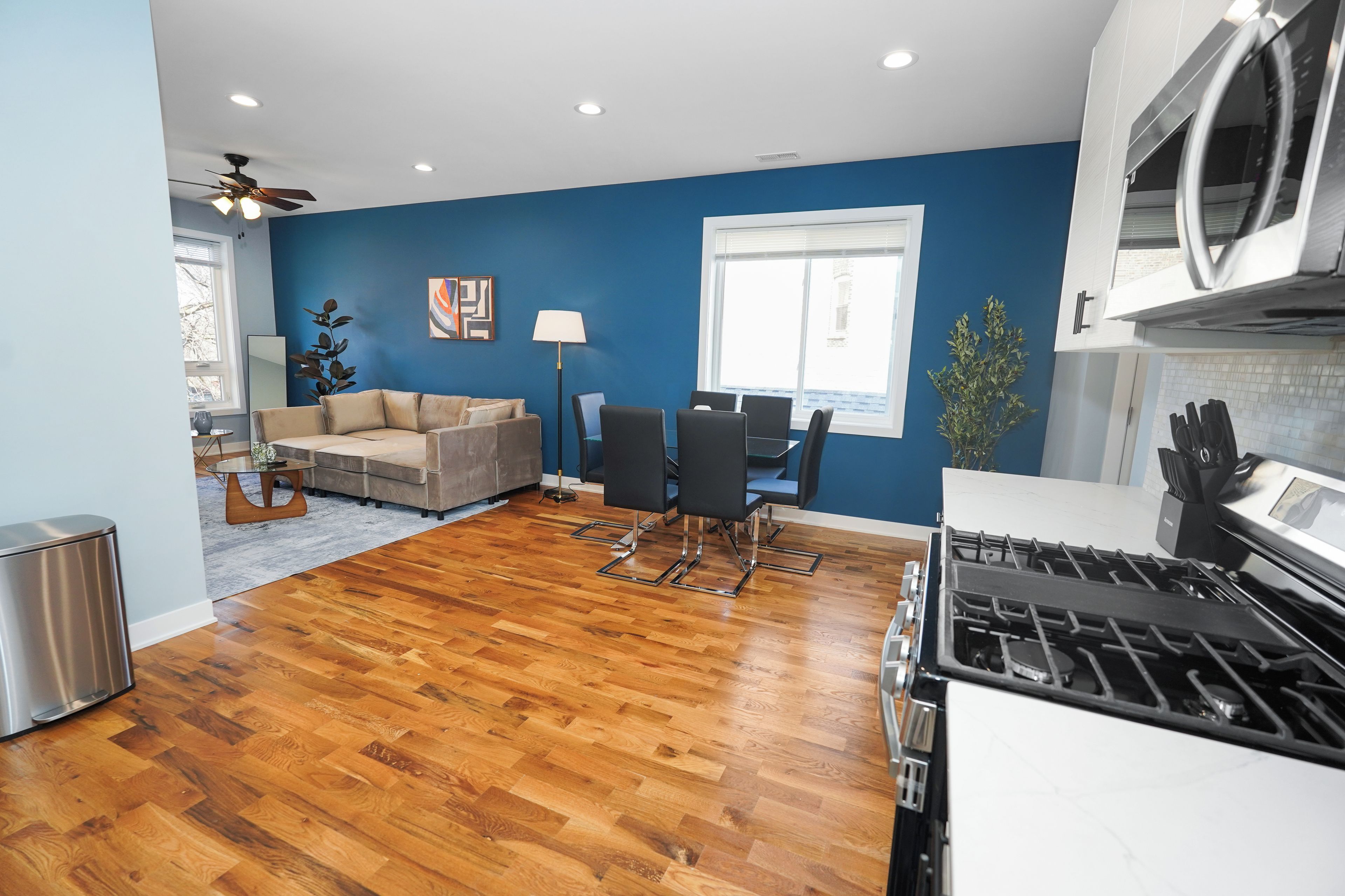 A modern kitchen with a stove, a dining area with a table and chairs, a sofa, and a ceiling fan.