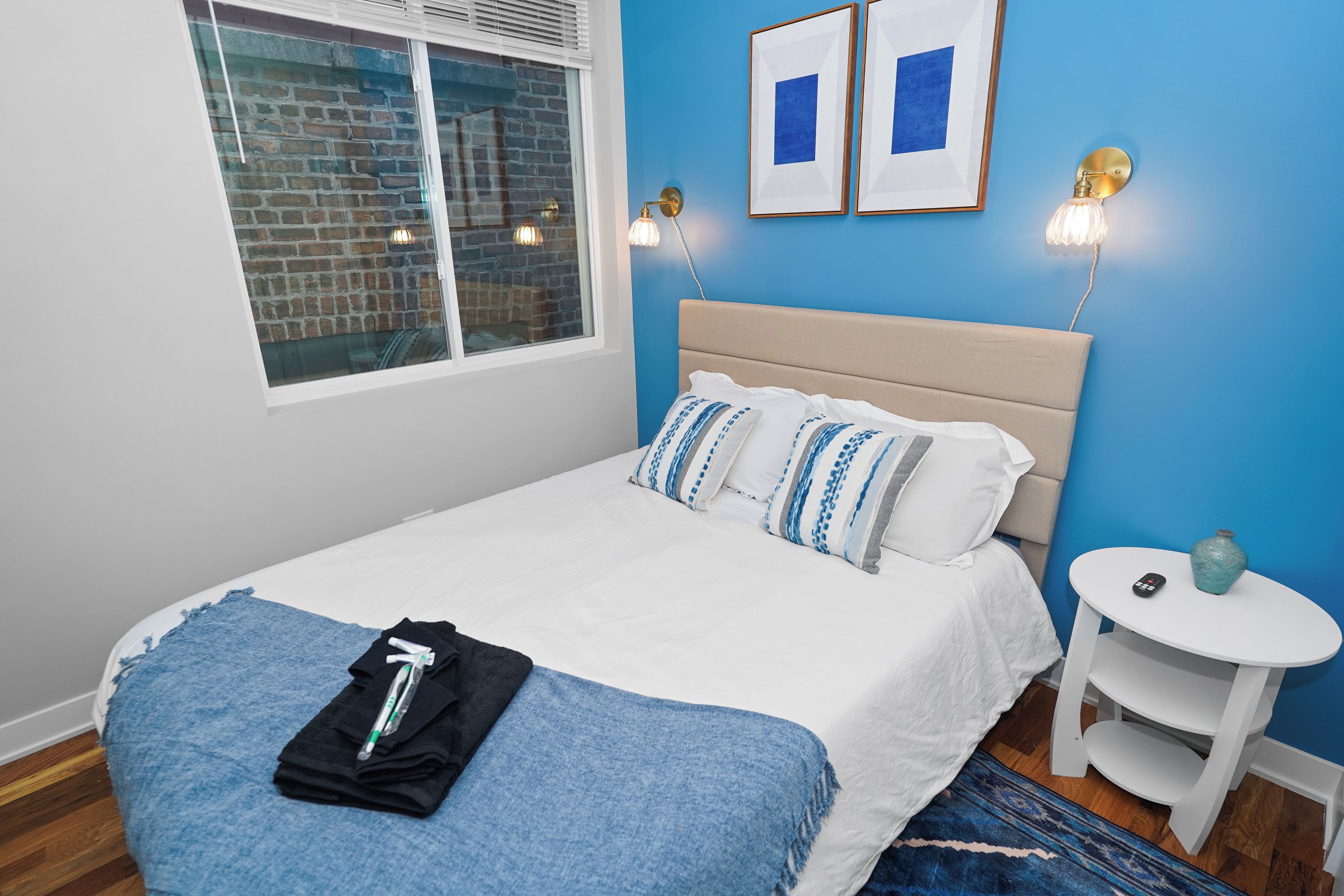 A neatly made bed with pillows, a bedside table with a vase, and a window with blinds.