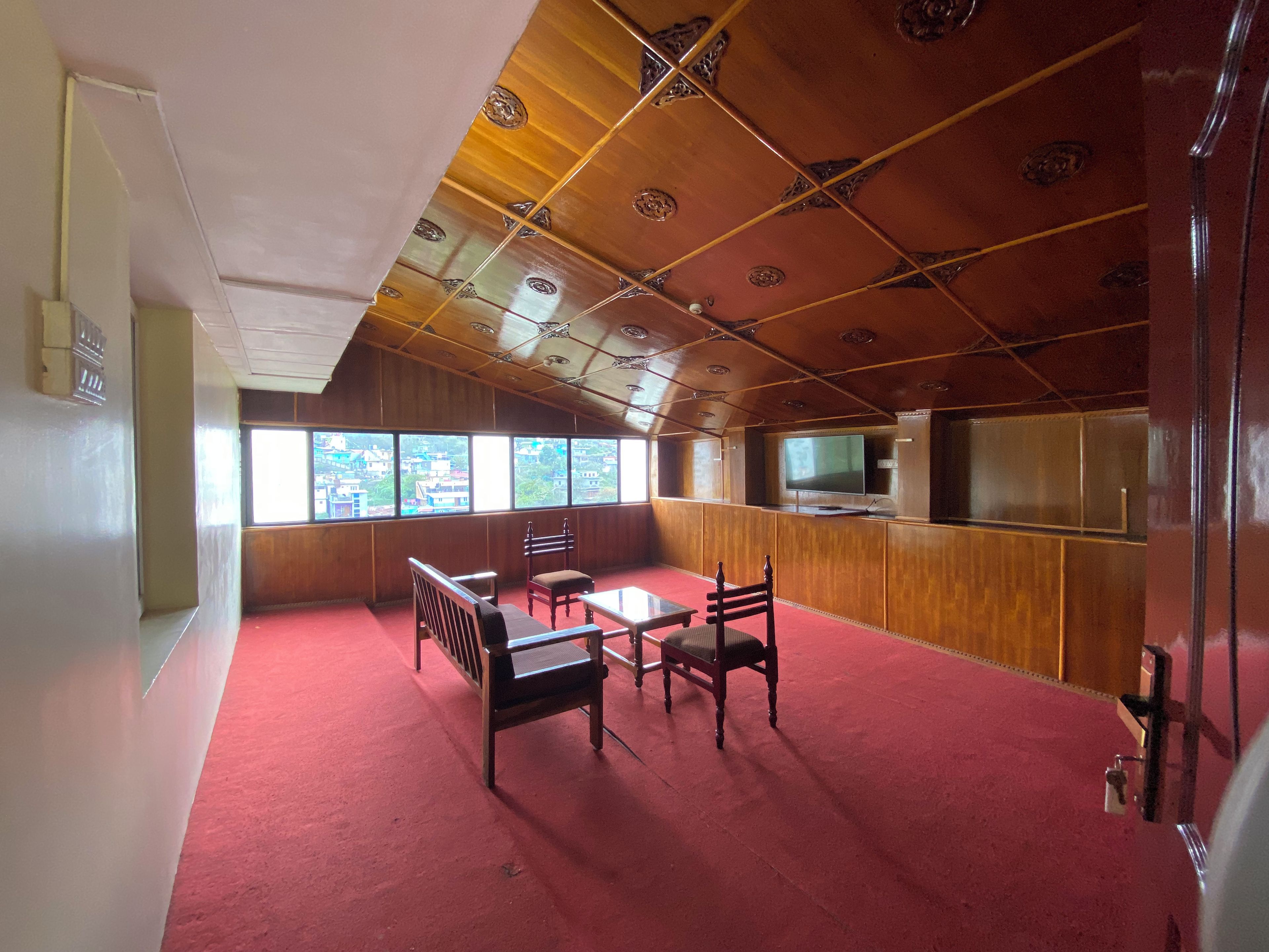 Una habitación con alfombra roja, techo de madera, un televisor y una ventana con vistas a edificios.