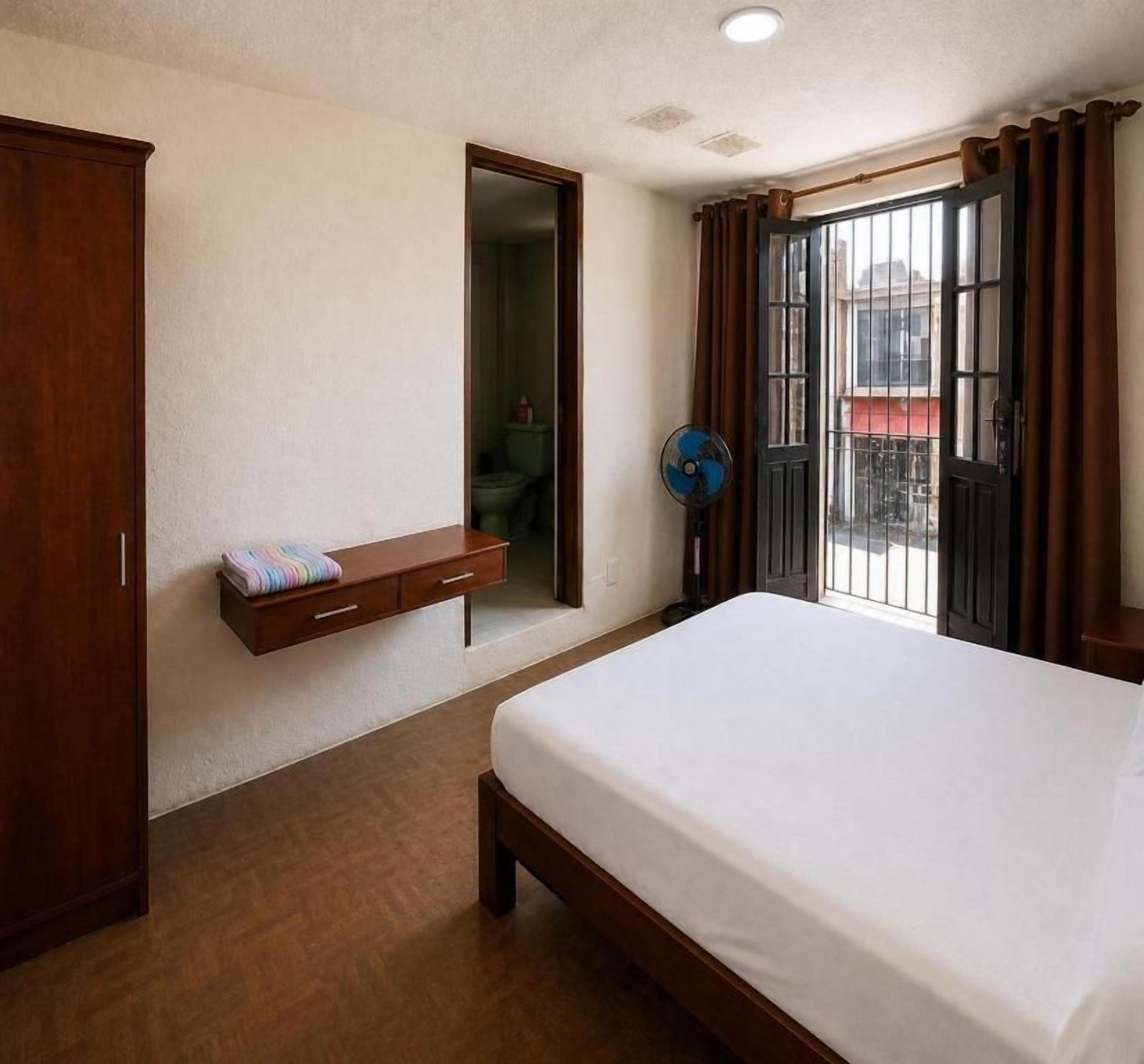 Un dormitori amb un llit, un armari de fusta, una petita prestatgeria, un ventilador i una porta que dona a una zona exterior.