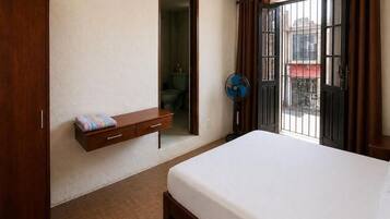 Un dormitori amb un llit, un armari de fusta, una petita prestatgeria, un ventilador i una porta que dona a una zona exterior.