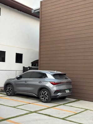 Un SUV gris estacionat a una entrada amb un edifici blanc i una paret marró al fons.