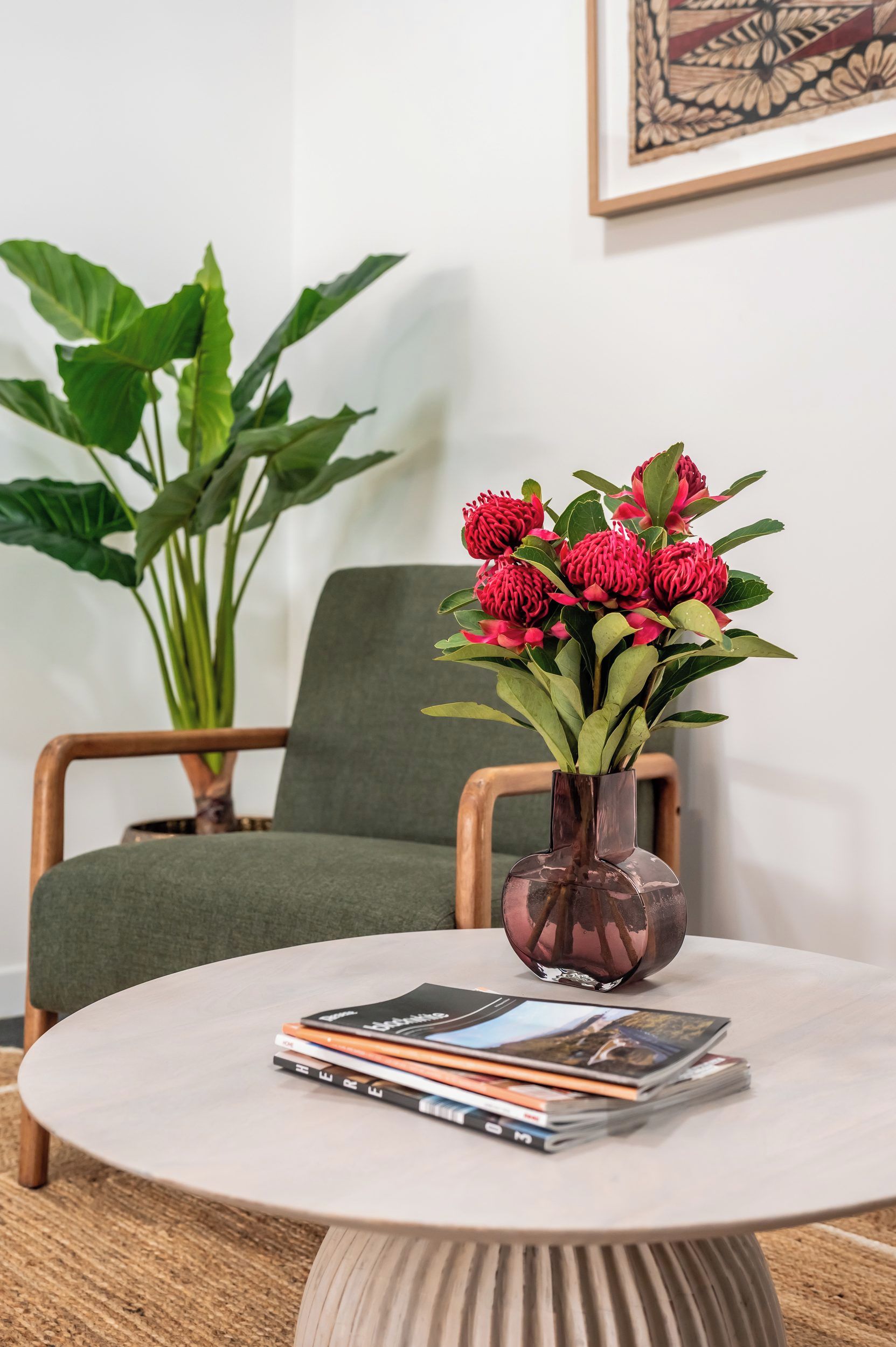 Taula rodona amb un ram de flors vermelles, una pila de revistes i una planta verda al fons.