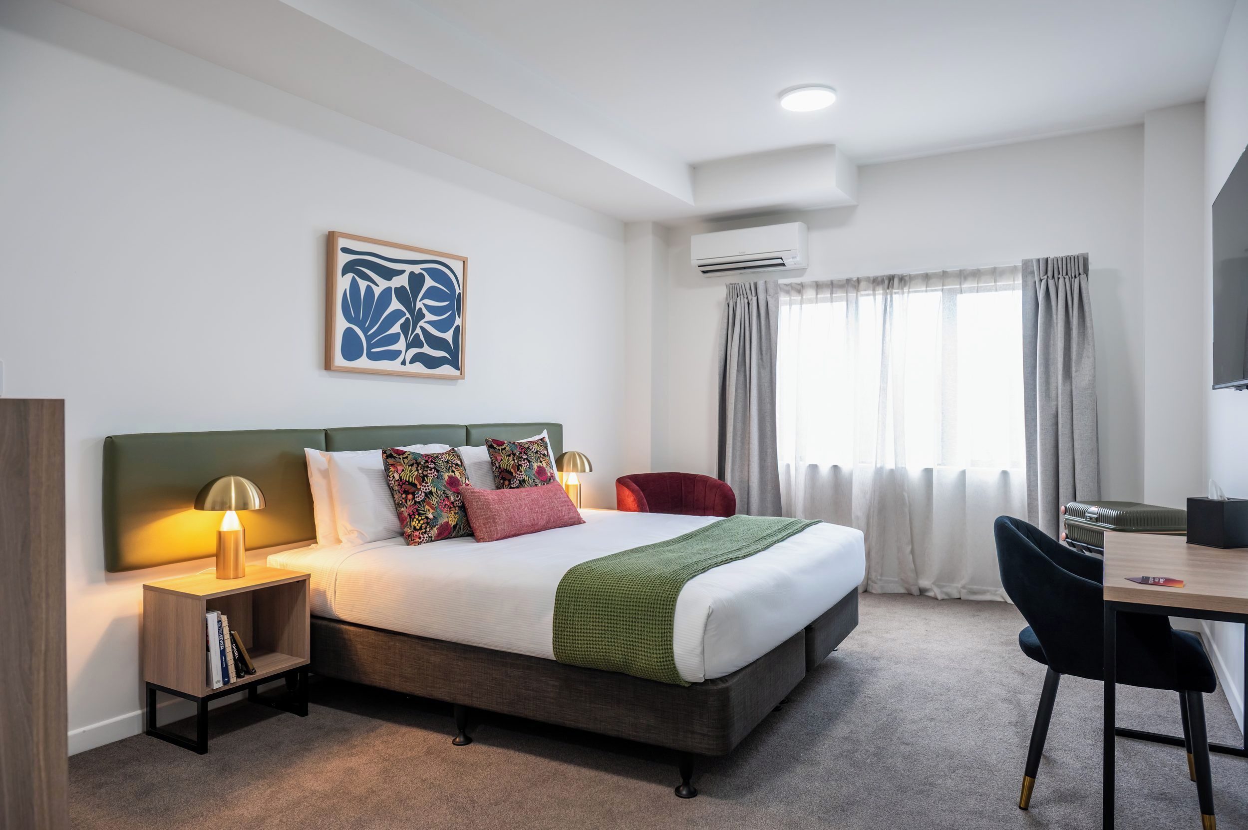 Une chambre d’hôtel comprenant un lit, des tables de chevet, un bureau, une chaise, un appareil de climatisation et une fenêtre avec des rideaux.