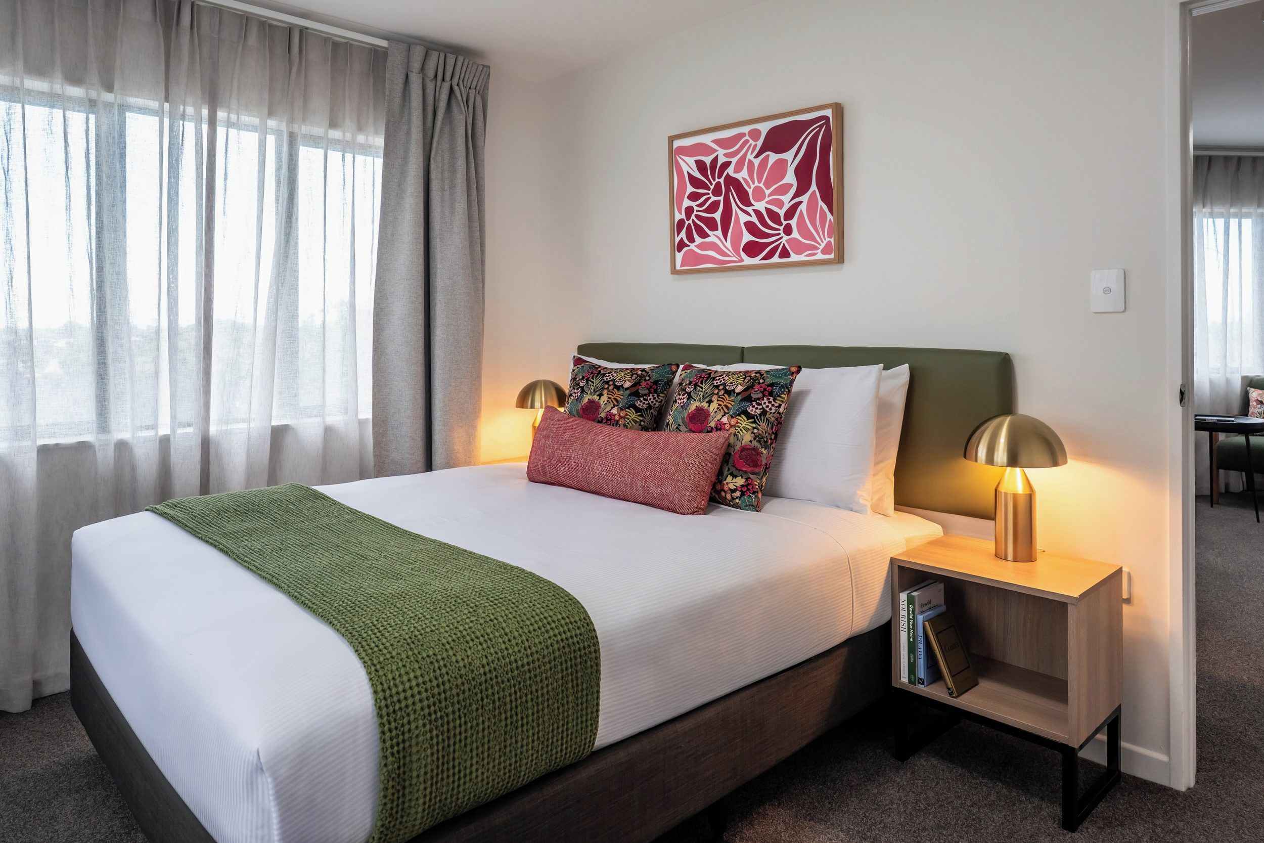 A hotel room with a bed, two bedside lamps, a green throw blanket, and a floral pillow.