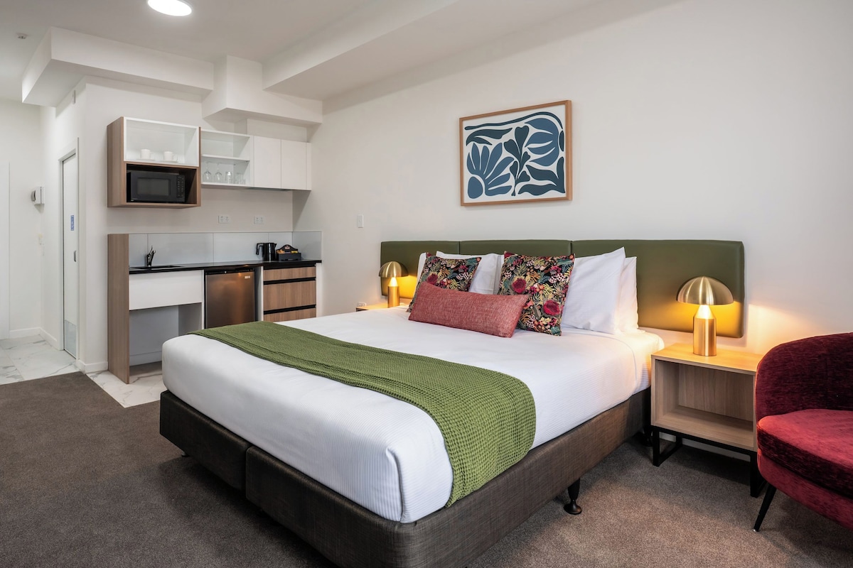 Una habitación de hotel con una cama, mesitas de noche, un escritorio, un microondas y una kitchenette.