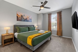 A hotel room with a bed, a ceiling fan, a painting, a window with curtains, and a flat-screen TV. - Stylish 2BR | Pool | Near Universal & Disney (Orlando)