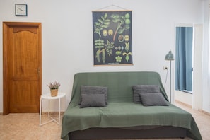 A room with a green sofa, a small side table with a plant, a poster of plants on the wall, a wooden door, and a window with curtains. - GuestReady - Canteras Surf, Relax & Work (Las Palmas de Gran Canaria)