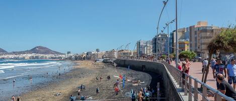 Eine Uferpromenade mit einem Steinweg, Strand und Stadtgebäuden im Hintergrund.