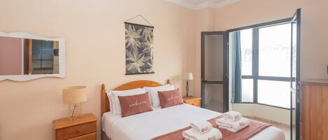 A bedroom with a bed, a nightstand, a mirror, a wall art piece, and a window.