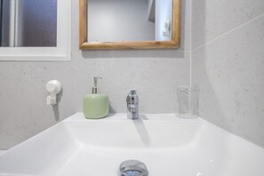 Un baño con un lavamanos blanco, un dispensador de jabón verde, un vaso de vidrio transparente y un espejo con marco de madera.
