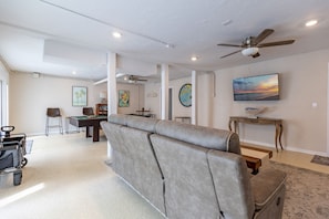 A spacious living room with a grey sofa, a pool table, a ceiling fan, and a television mounted on the wall. - Sea Cove Castle by Duncan Real Estate (Anna Maria)