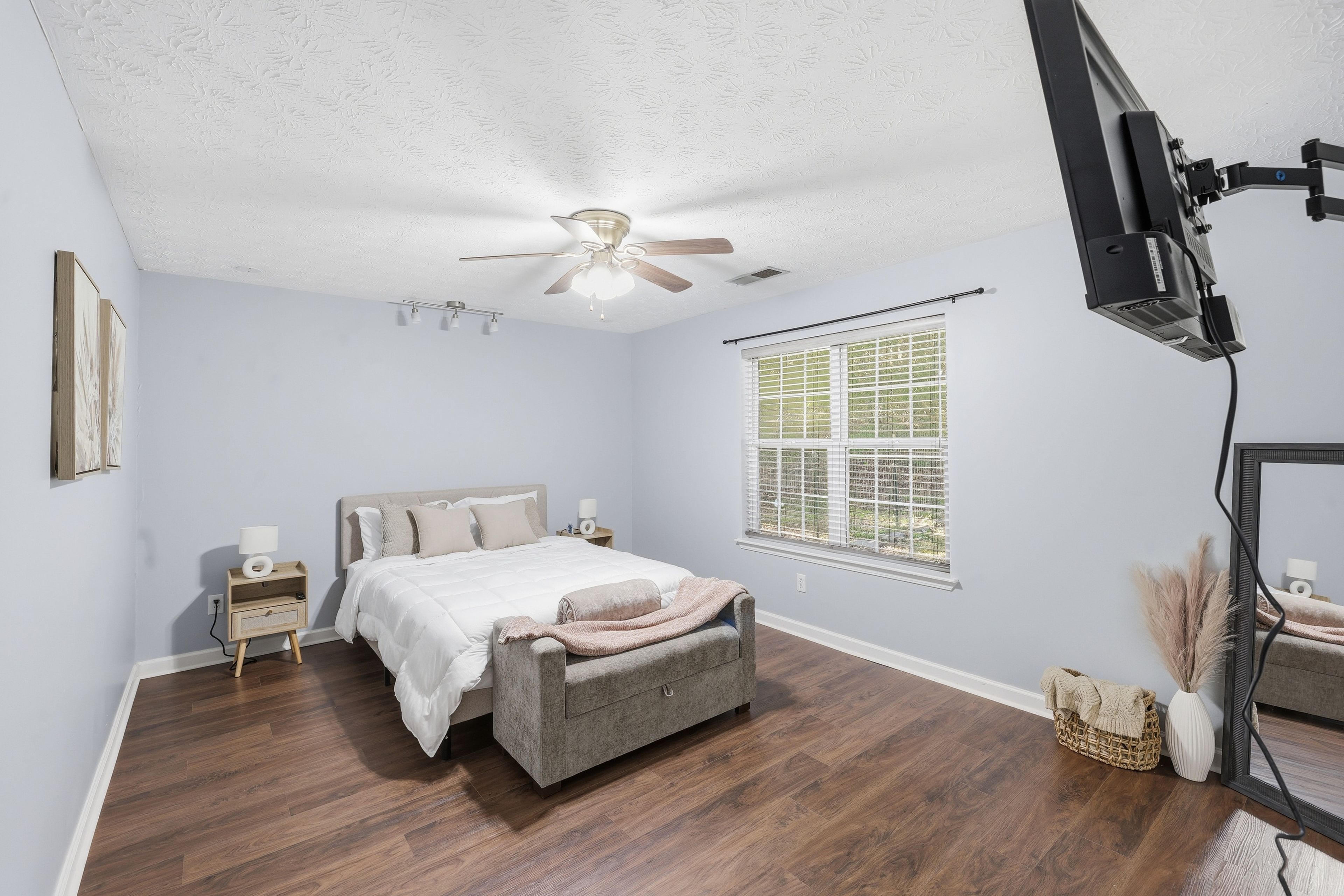 A bedroom with a bed, nightstands, a ceiling fan, a window with blinds, and a television mounted on the wall.