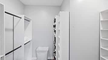 Um corredor estreito com estantes brancas, um cesto de lixo e um ventilador preto.