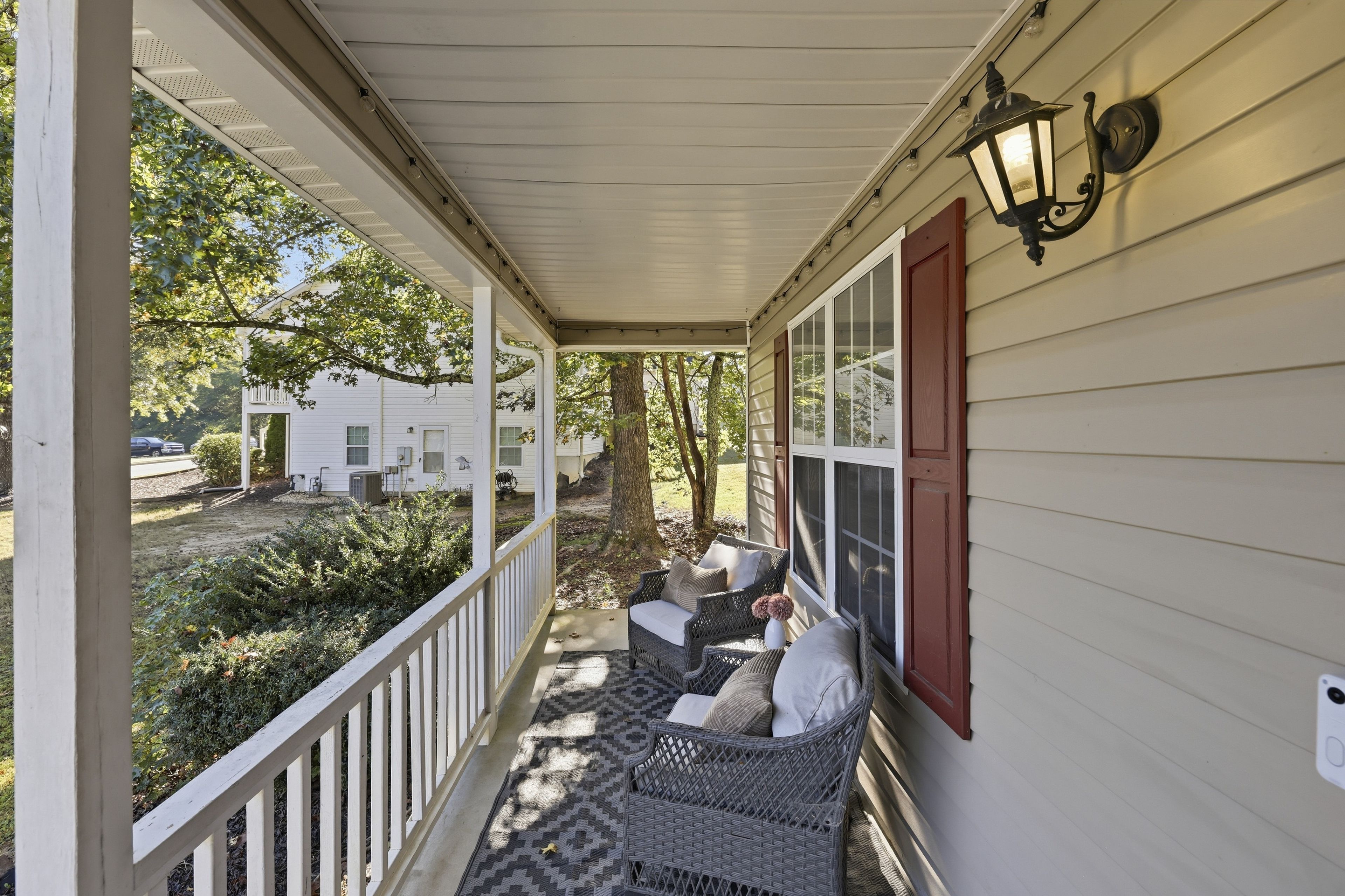 A covered porch with wicker furniture and a red door, overlooking a yard with trees and a white house.