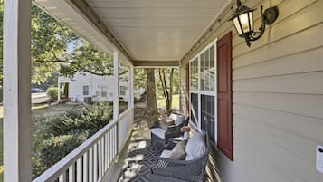 A covered porch with wicker furniture and a red door, overlooking a yard with trees and a white house.