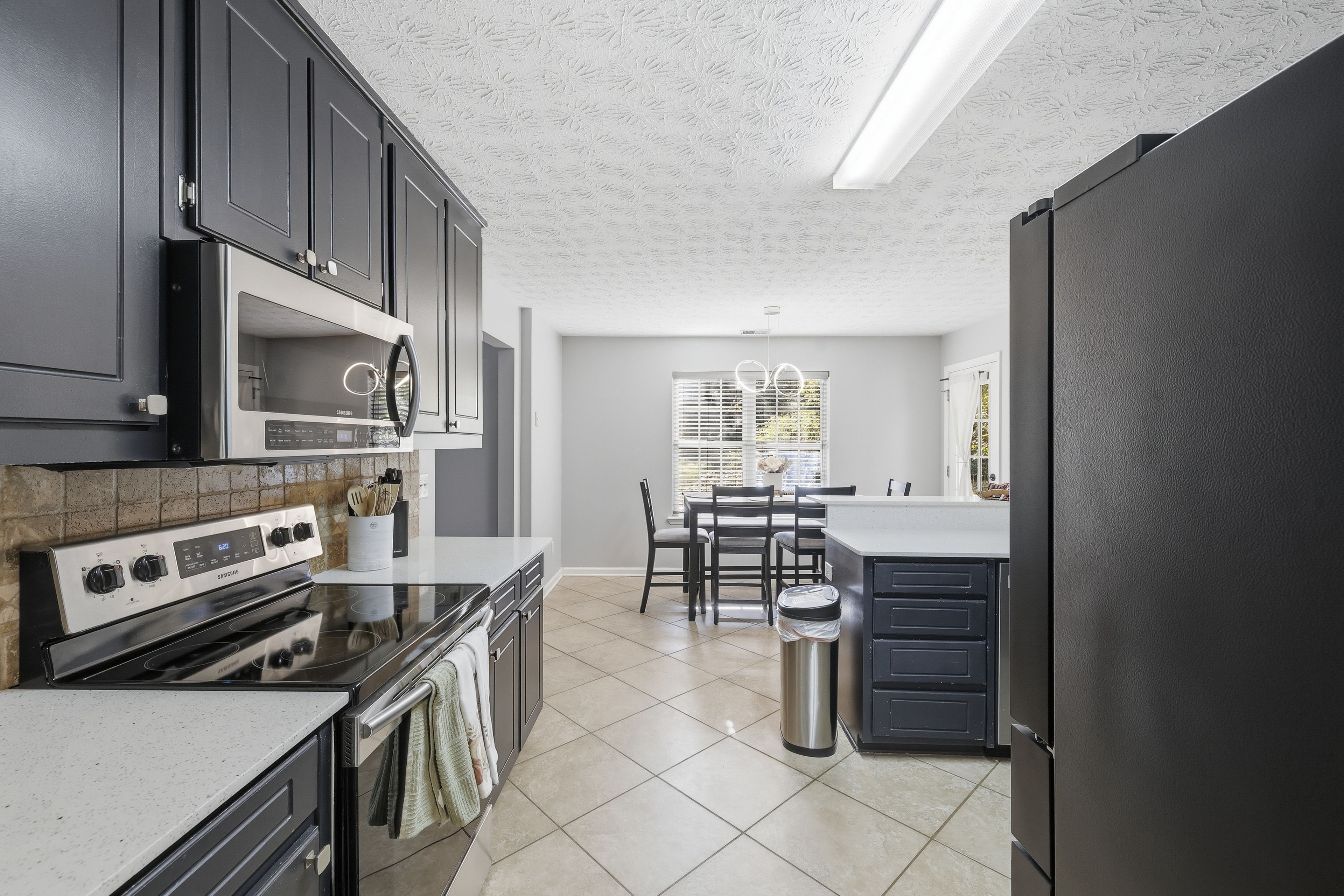 A modern kitchen with a built-in oven, microwave, and stainless steel appliances. There is a dining area with a table and chairs, and a trash can.