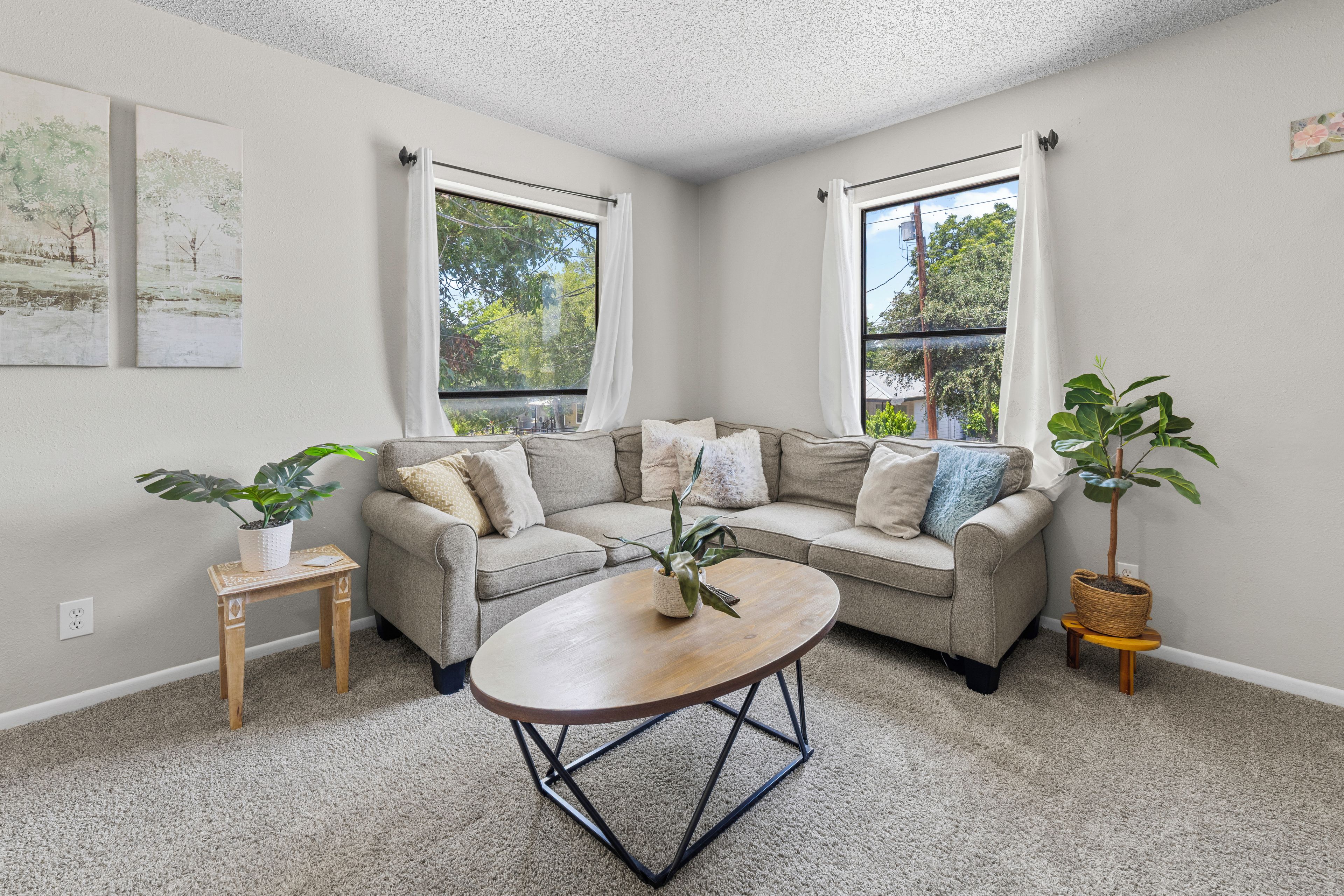 A corner living room with a beige sofa, two windows with white curtains, a small wooden table, and a coffee table.