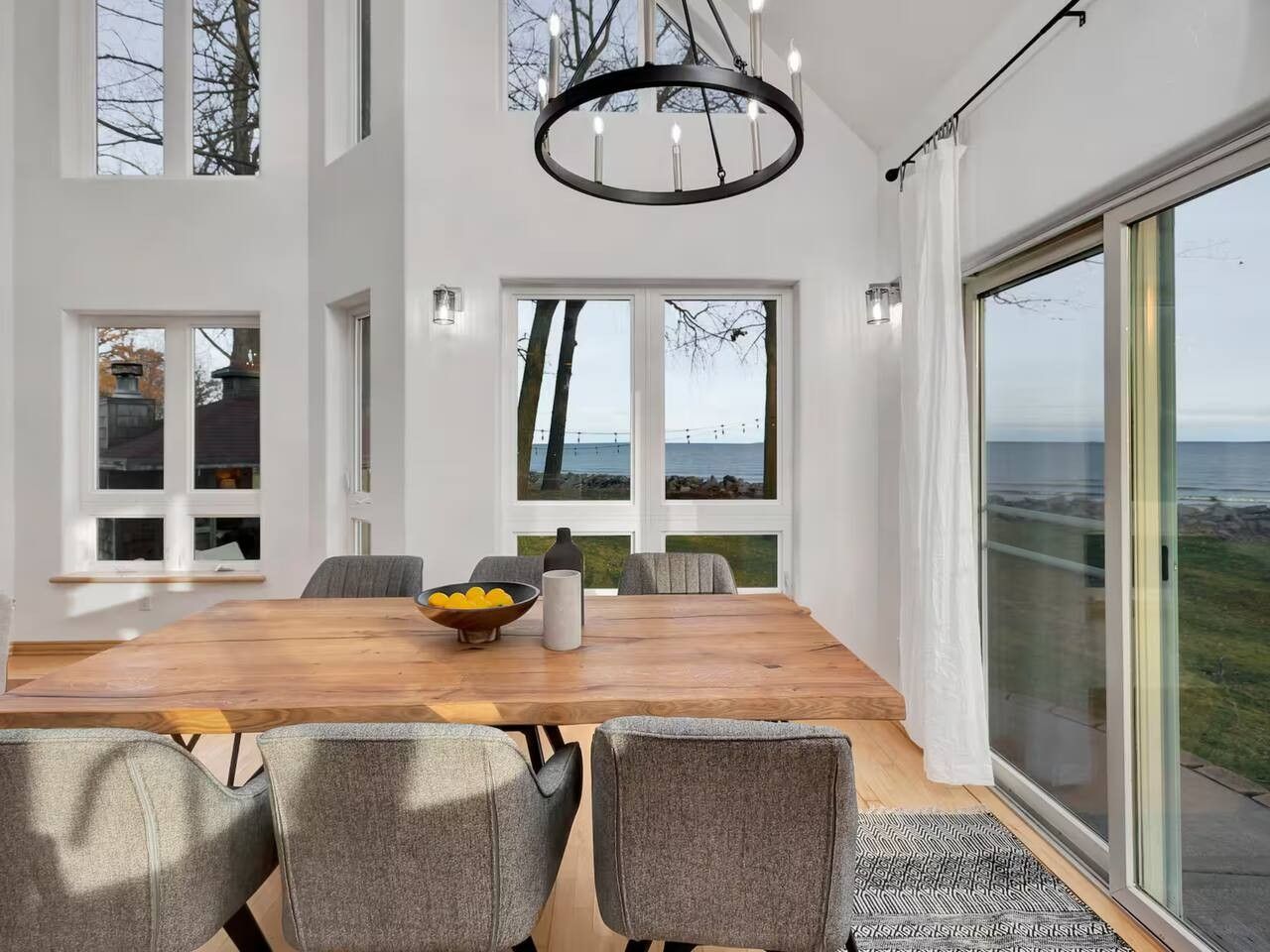 Ett modernt matområde med ett träbord, grå klädda stolar, en kristallkrona och stora fönster med utsikt över havet.