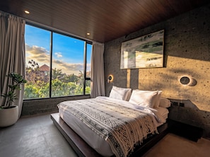 Un dormitorio con un ventanal grande, una cama con ropa de cama estampada, una planta en maceta y un aplique de pared.