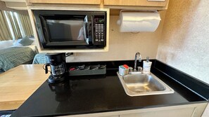 Een compact keukenhoekje met een magnetron, koffiezetapparaat, spoelbak en een klein kastje.