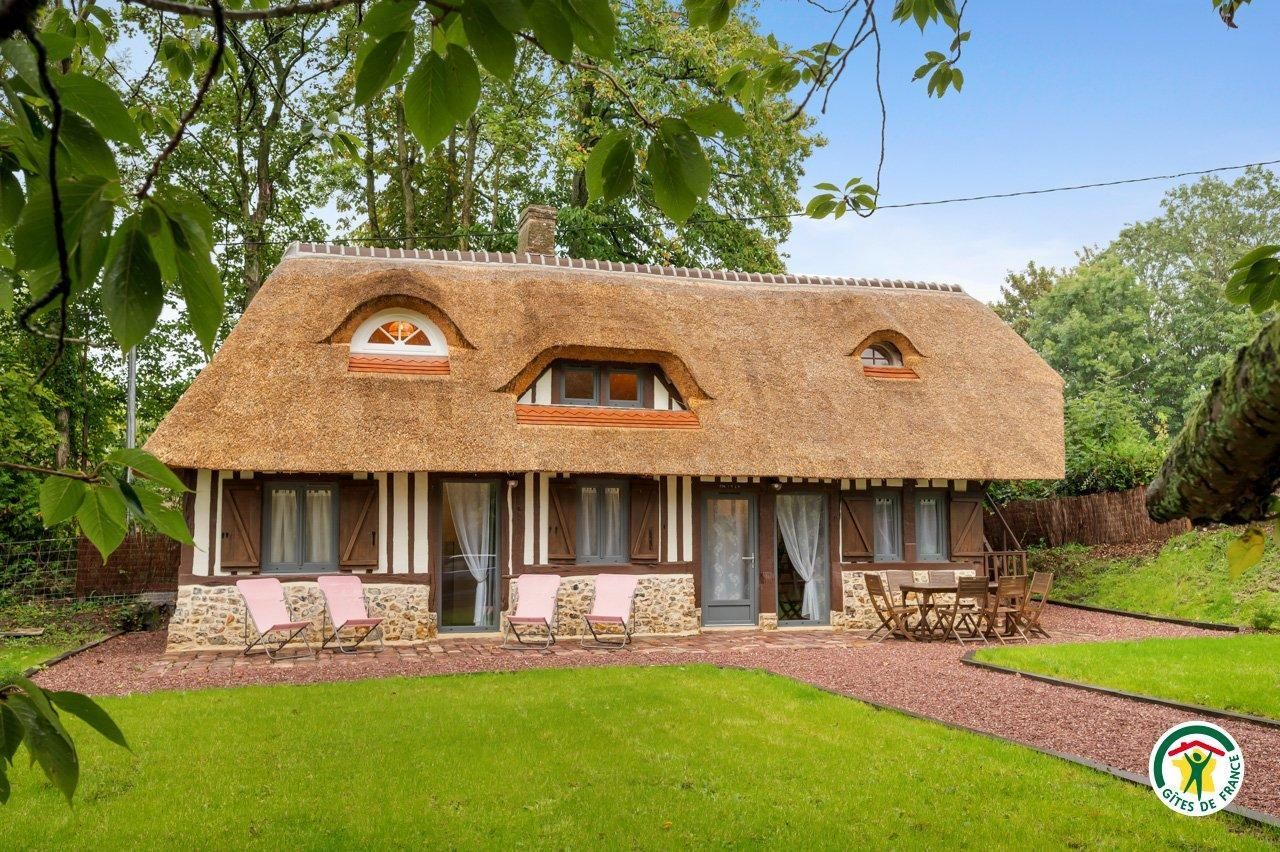 Une maison rustique avec un toit de chaume, des murs en pierre et un petit porche à l’avant, agrémenté de deux chaises.