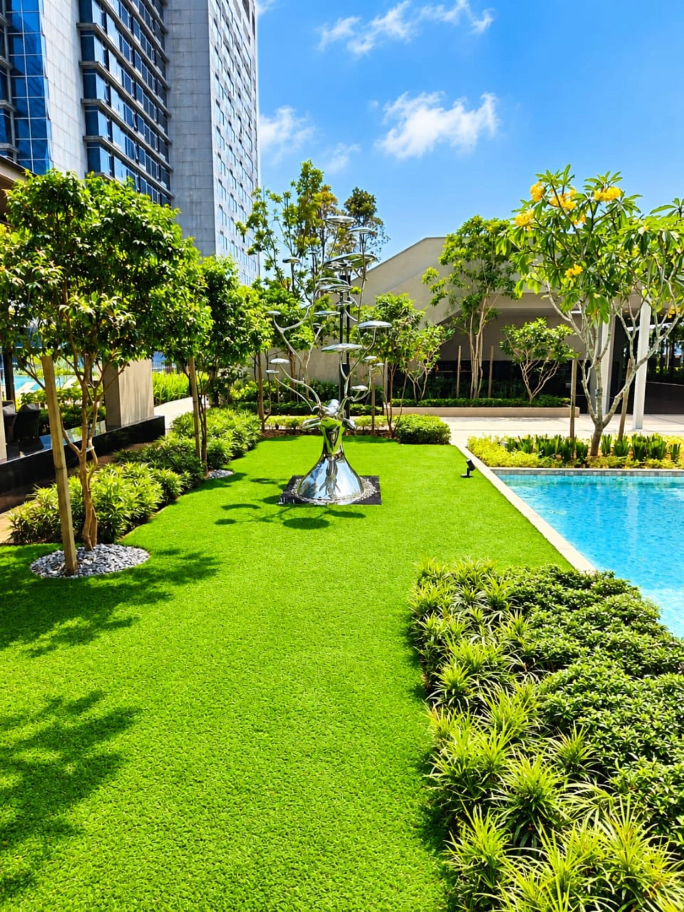 A landscaped garden with a central sculpture, surrounded by well-maintained grass and trees, and a pool on one side.
