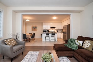 A modern living room with a sofa, a coffee table, and a dining area with chairs and a table. - Niagara Falls Family Retreat - Sleeps 8 Group (Thorold)