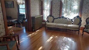 A room with a wooden floor, a vintage sofa, a chair, a radiator, and patterned wallpaper.