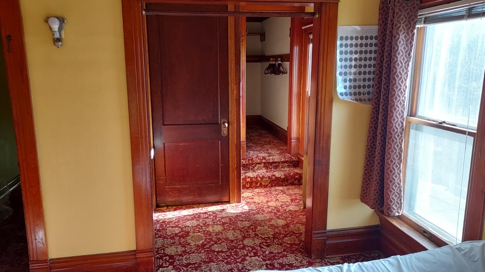 A room with a wooden door, a patterned carpet, a window with curtains, and a wall-mounted light fixture.
