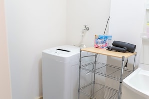 A laundry area with a washing machine, a drying rack, and a shelf with a hairdryer and a box of laundry detergent. - Ocean Nishikujo (Osaka)
