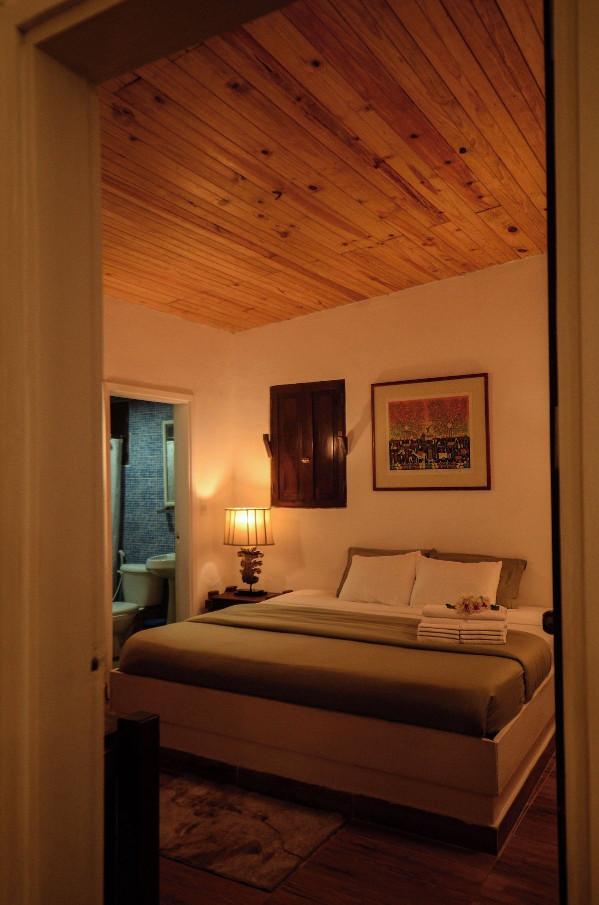 Un dormitorio con techo de madera, una cama con almohadas, una mesita de noche con lámpara, un cuadro en la pared y una ventana con vistas al exterior.