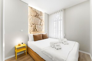 Hotellihuone, jossa on sänky, yöpöytä, kivimäistä pintaa jäljittelevä seinä ja ikkuna, jossa on läpikuultavat verhot.