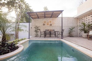 Ein Schwimmbecken mit überdachter Terrasse, umgeben von Pflanzen und einer weißen Mauer mit dekorativen Platten.