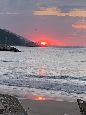 Escena de playa con un atardecer sobre el agua, olas y el horizonte de una ciudad distante.
