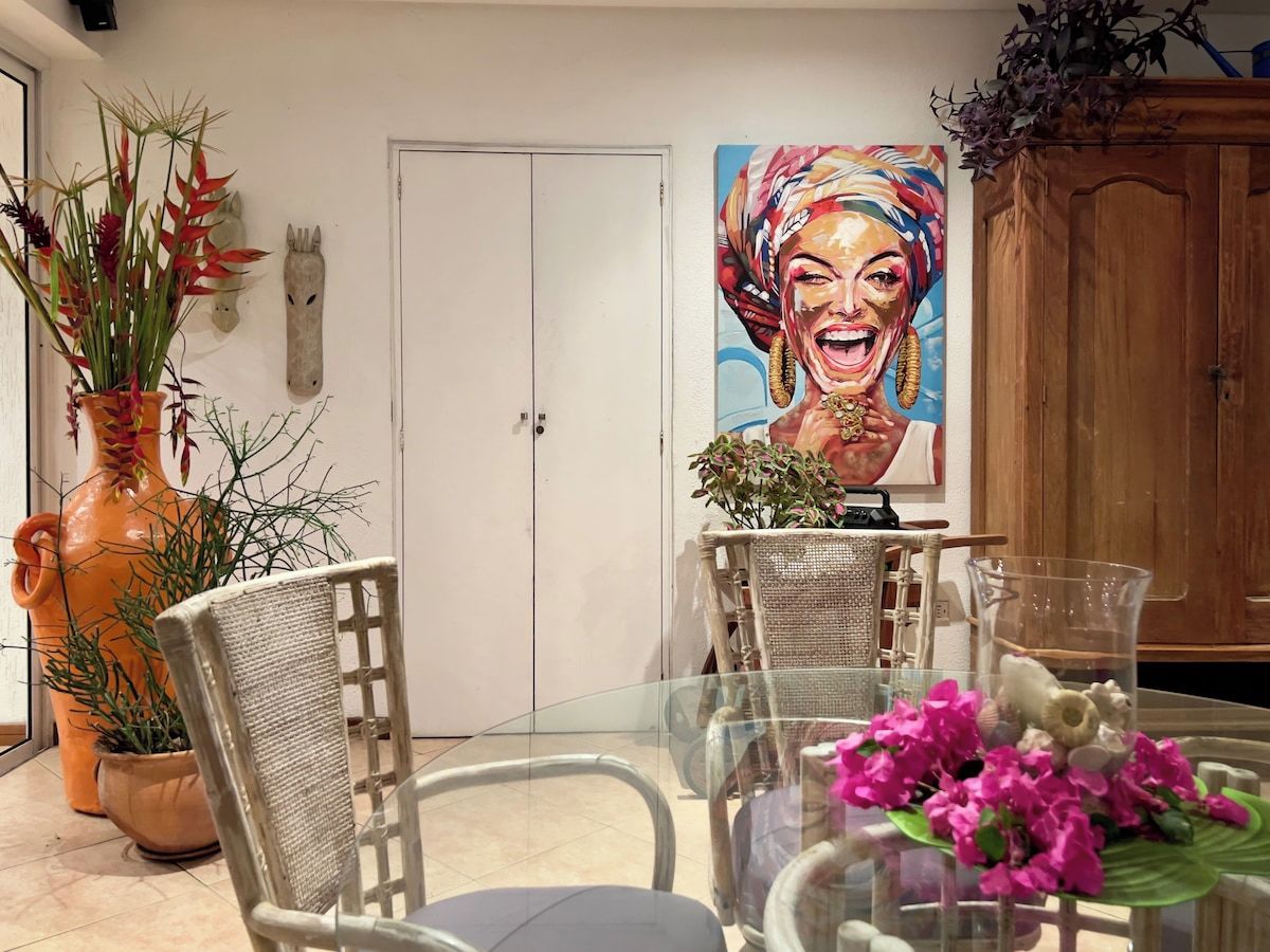 A dining area with a glass table, wicker chairs, and a large painting on the wall.