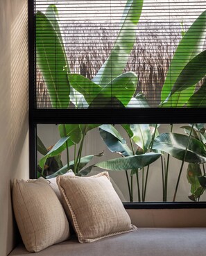 Una ventana con vistas a grandes hojas verdes, una pared con textura y un banco acolchado con dos almohadas.