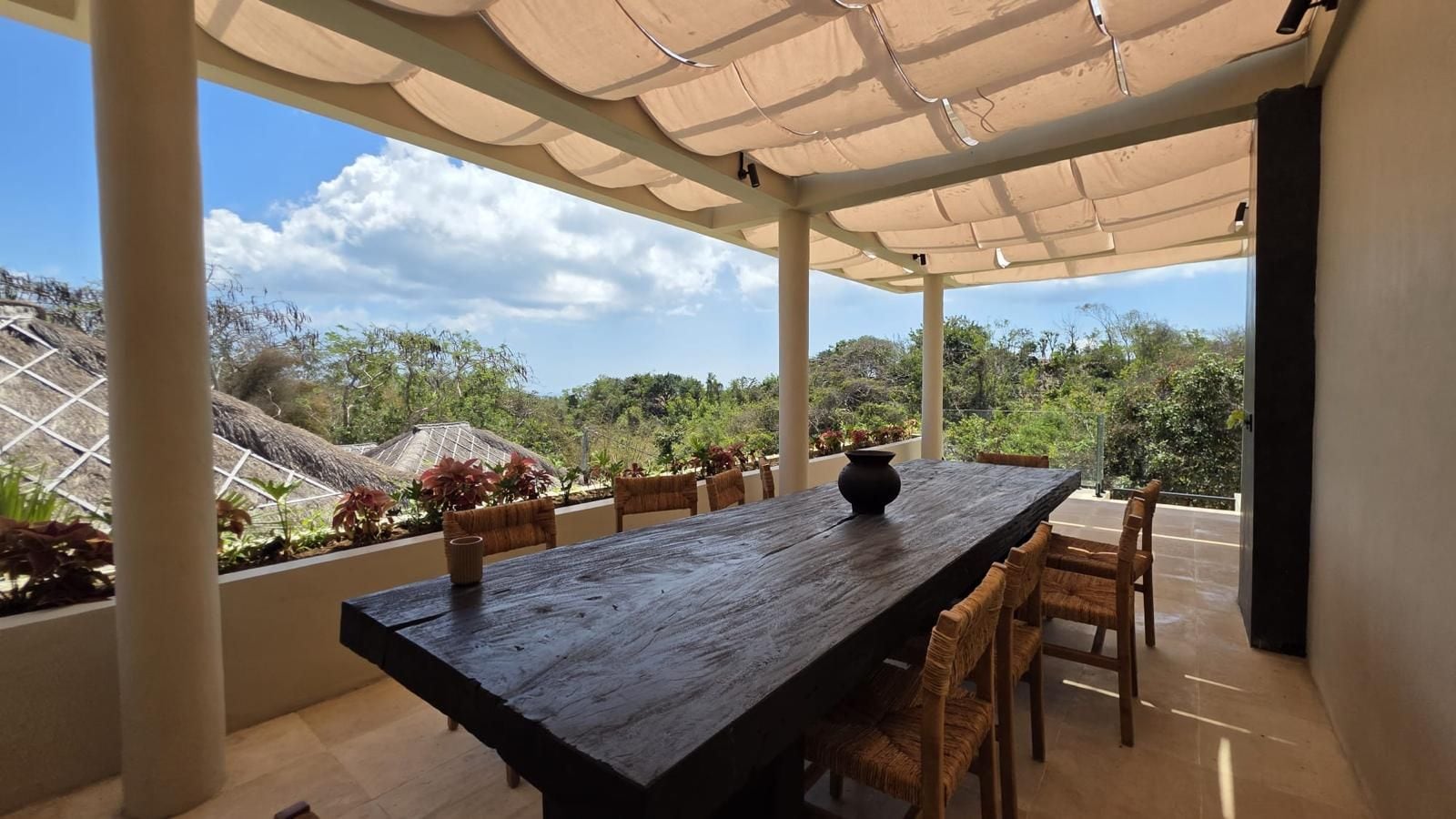 Pátio externo com mesa e cadeiras de madeira, cercado por plantas e vista para a paisagem.