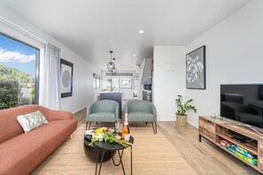 A modern living room with a sofa, two armchairs, a coffee table with flowers and a bottle, a TV, and a kitchen in the background. - Soleil by the Sea (Whitianga)