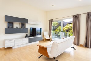 A modern living room with a flat-screen TV, a sofa, and a chair. There is a balcony with curtains and a view of palm trees. - Villa with a pool and ocean views in Santa Úrsula; spacious villa that sleeps up to 10 people (Santa Úrsula)