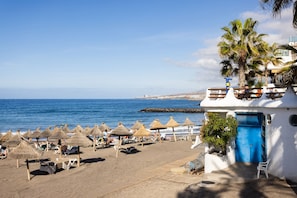 A beach with thatched umbrellas, lounge chairs, and a building with a blue door. - Apartment in a prime location with a terrace, pool, and the beach just a few steps away (Costa Adeje)