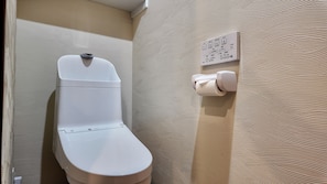 A toilet with a white seat and lid, a wall-mounted flush button, and a toilet paper holder. - JIA Kamei Osaka (Osaka)