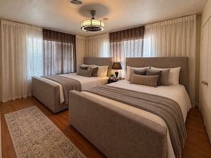 A hotel room with two beds, a ceiling fan, a nightstand with a lamp, and a large window with curtains.
