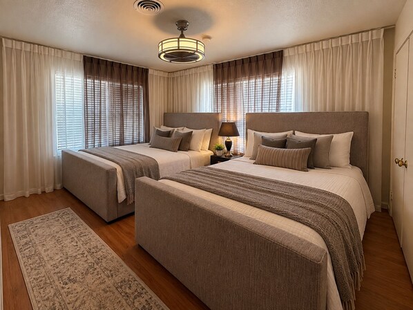 A hotel room with two beds, a ceiling fan, a nightstand with a lamp, and a large window with curtains.