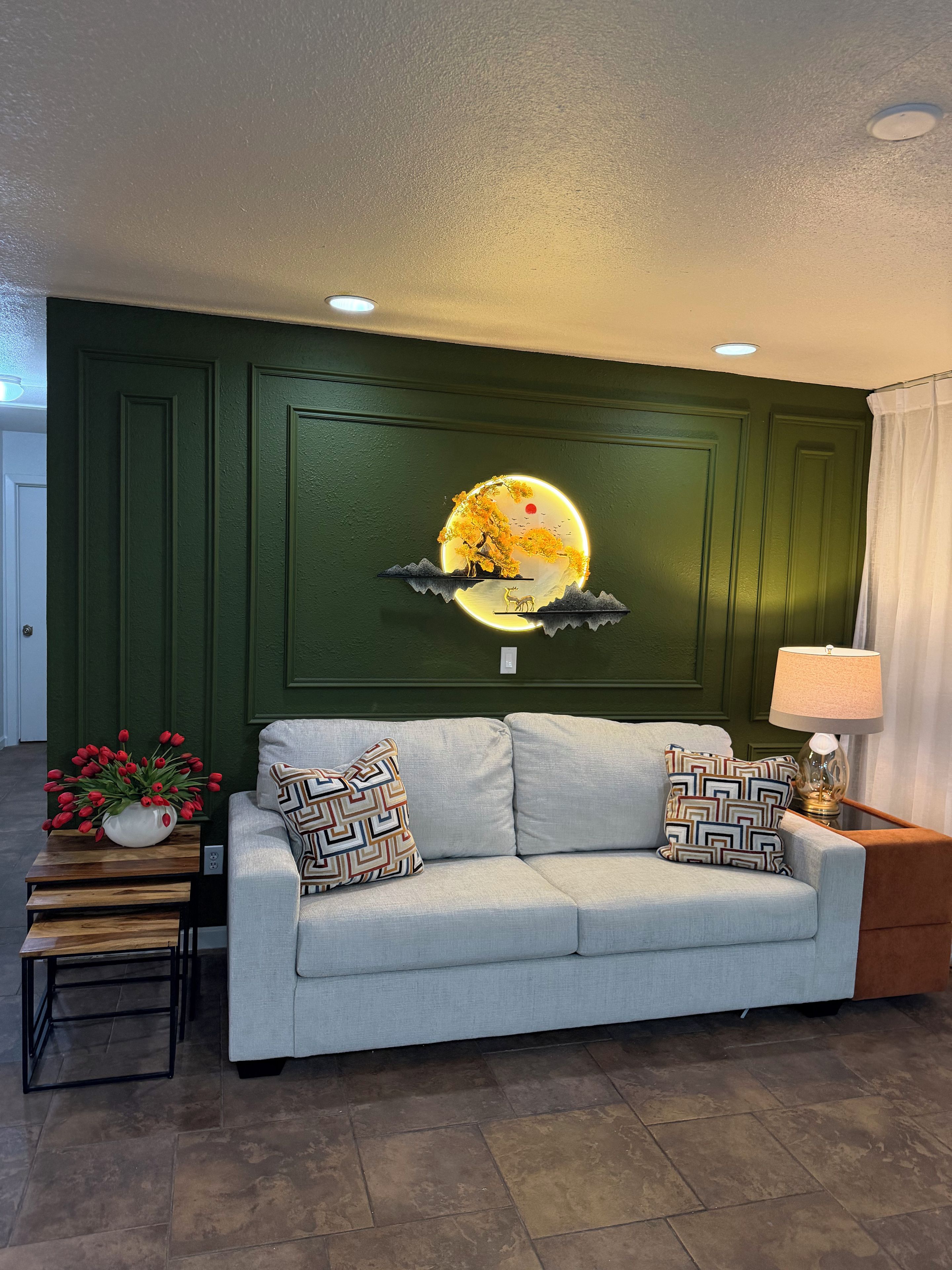A living room with a green wall, a sofa with patterned cushions, a small table with a vase of red flowers, and a wall-mounted artwork of a tree and moon.