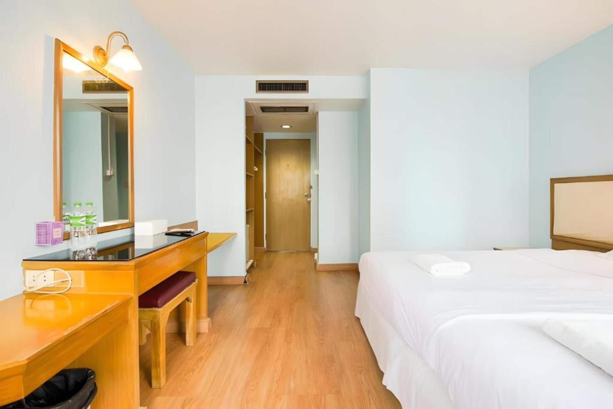 A hotel room with a wooden desk, a large mirror, a bed, and a door leading to another room.
