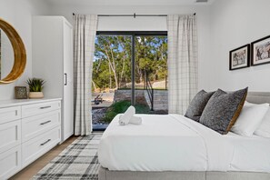 Ein Schlafzimmer mit Bett, Schminktisch, Spiegel und Blick auf einen Außenbereich mit Bäumen und einer Steinmauer.