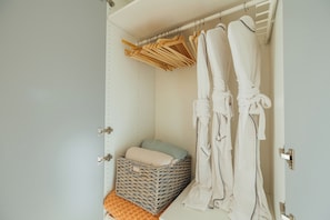 Ein begehbarer Kleiderschrank mit hängenden Roben, einem Korbwäschekorb und einem Bett mit blauem Kopfkissen.